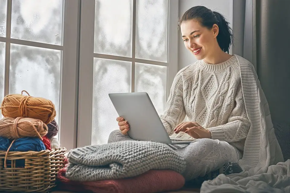 Happy beautiful woman watching videos or enjoying entertainment content on a laptop sitting near windows in the house in the winter.