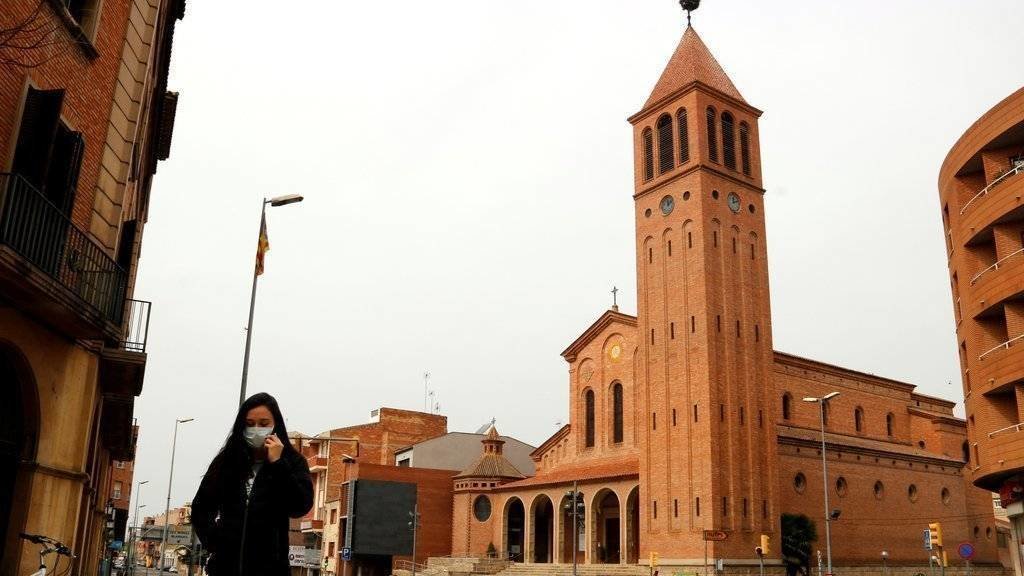 Pla general on es pot veure el campanar de la parròquia de Sant Jaume de Mollerussa i una noia caminant pel carrer amb mascareta per evitar contagis del coronavirus, el 18 de març de 2020. (Horitzontal)