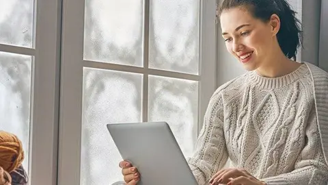 Happy beautiful woman watching videos or enjoying entertainment content on a laptop sitting near windows in the house in the winter.