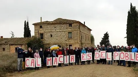 Pla general d'alguns dels participats a l'acte Vallmanya amb un cartell que reclama que "Cal Macià és del poble" i la casa al fons, l'11 d'abril de 2021. (Horitzontal)