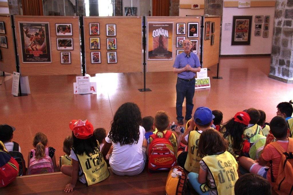 Pla general on es veuen a diversos nens i nenes del casal d'estiu de la Seu d'Urgell escoltant les explicacions del cinèfil i col·leccionista Josep Benavent sobre l'exposició 'Herois i superherois'. Imatge del 6 d'agost de 2019 (Horitzontal).