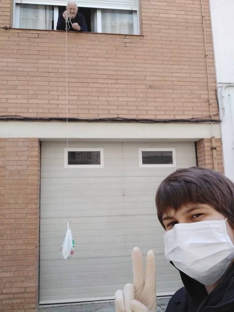 Pla general on es veu un dels usuaris del servei Ajude(M)ollerussa rebent a domicili la compra gràcies a un dels voluntaris, que es veu en primer terme. Imatge facilitada per l'Ajuntament de Mollerussa el 27 de març de 2020 (Vertical).