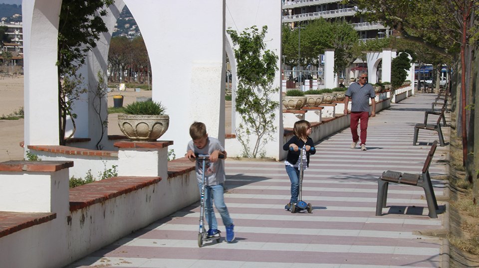 Pla obert del Passeig de Palamós amb dos nens amb un patinet i el seu avi al darrere aquest diumenge 26 d'abril de 2020. (Horitzontal)