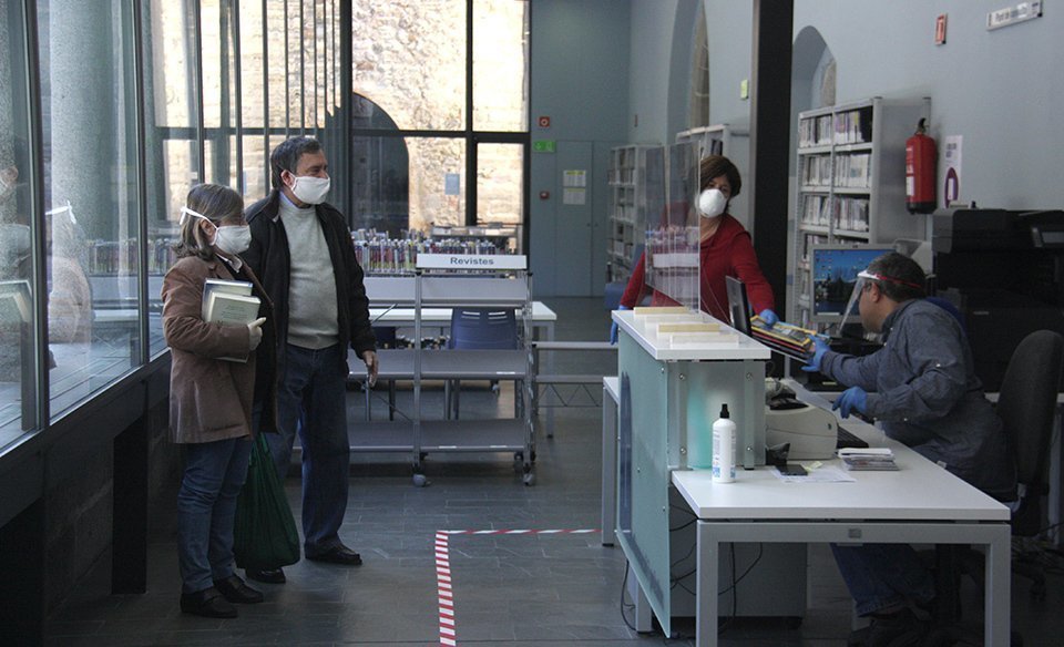 Pla general de l'interior de la Biblioteca Comtat de Cerdanya de Puigcerdà on es veuen diversos usuaris amb mascareta i separats dels treballadors a punt d'endur-se nous llibres en préstec. Imatge del 12 de maig de 2020 (Horitzontal).