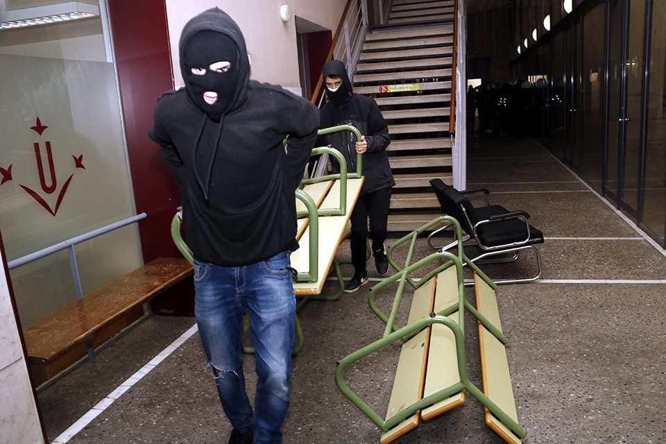 Pla mitjà on es poden veure dos joves encaputxats i amb la cara tapada traslladant un banc per fer barricades i reforçar la tancada a la UdL per Pablo Hasél, el 15 de febrer de 2021. (Horitzontal)