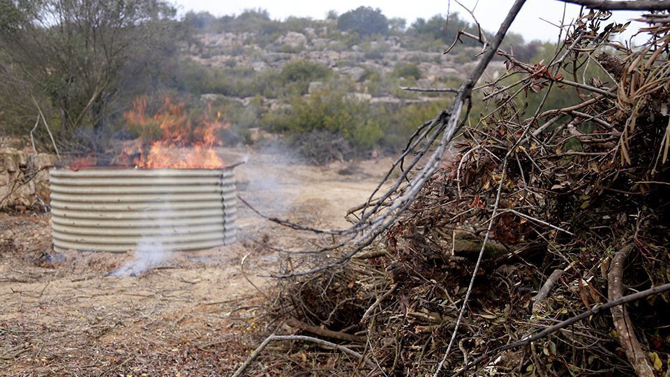 Pla tancat de les restes de poda que s'han utilitzat per fer biocarbó en una finca de Torrebesses i al fons, el forn per elaborar-lo, el 24 de desembre de 2021. (Horitzontal)